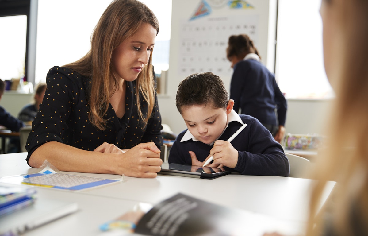 Young female teacher working with a Down syndrome schoolboy sitting at desk using a tablet computer and stylus in a primary school classroom